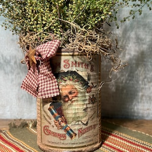 May include: A vintage-style Christmas decoration featuring a cylindrical container adorned with a Santa Claus image and the text "Tom Smith's Christmas Novelties". The container is filled with dried greenery and accented with a red and white checkered bow and a star.