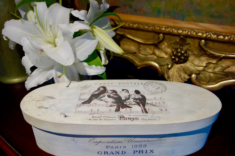 May include: An oval wooden box with a distressed white finish. The lid features a graphic of four birds perched on musical notes, the text "CARTE POSTALE," and a Paris postmark. The side displays the text "Exposition Universelle PARIS 1889 GRAND PRIX."
