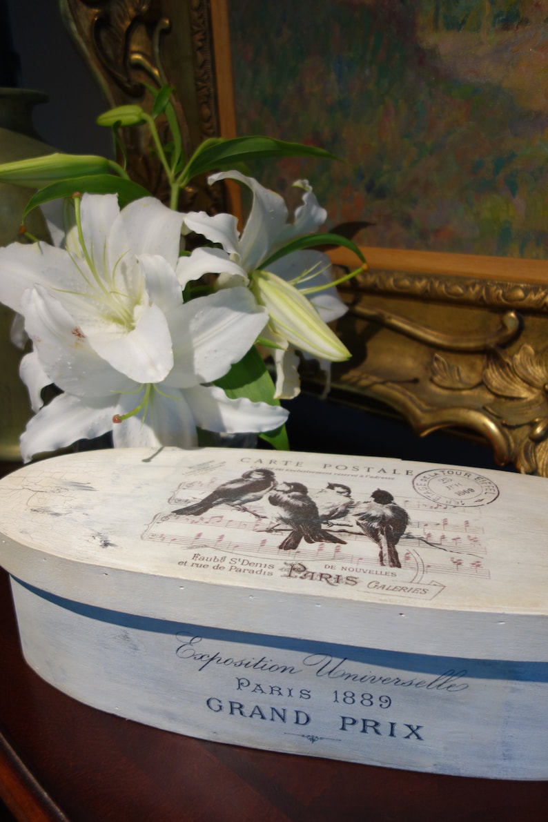 May include: An oval wooden box with a distressed white finish. The lid features a graphic of four birds on a musical staff with the text CARTE POSTALE and PARIS. The side displays the text Exposition Universelle PARIS 1889 GRAND PRIX.