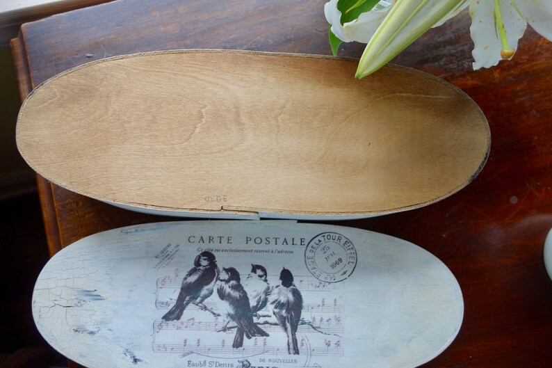 May include: An oval wooden box with a removable lid. The lid features a white distressed finish with a graphic print of four birds perched on musical notes, the text "CARTE POSTALE," and a circular "TOUR EIFFEL" postmark stamp.