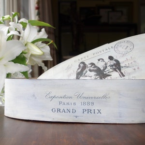 May include: An oval wooden box with a distressed white finish. The front features the text Exposition Universelle Paris 1889 GRAND PRIX. The lid displays a graphic of birds and the text CARTE POSTALE with a circular postmark stamp.