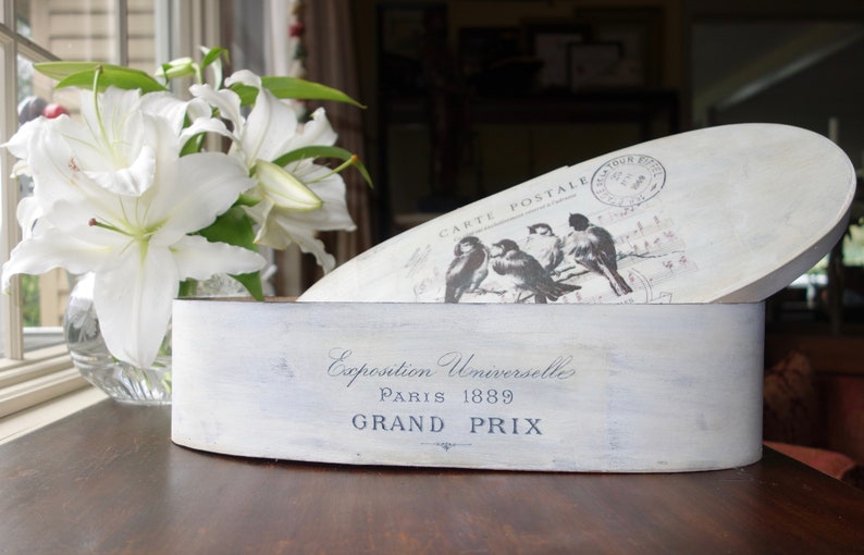 May include: An oval wooden box with a distressed white finish. The front features the text Exposition Universelle Paris 1889 GRAND PRIX. The lid displays a graphic of birds and the text CARTE POSTALE with a circular postmark stamp.