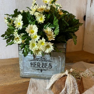 May include: A galvanized metal planter with the word "HERBES" printed on the front, filled with a bouquet of white daisies.