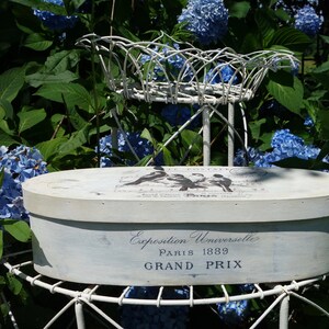 May include: An oval, off-white wooden box with a distressed finish, featuring black bird illustrations and the text "Exposition Universelle PARIS 1889 GRAND PRIX" printed on the side and lid, resting on a white metal wire stand.