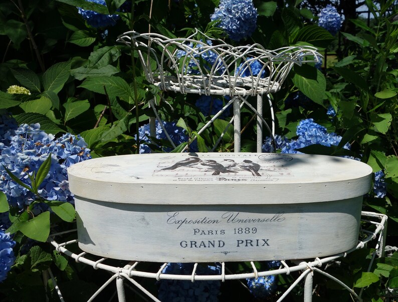 May include: An oval, off-white wooden box with a distressed finish, featuring black bird illustrations and the text "Exposition Universelle PARIS 1889 GRAND PRIX" printed on the side and lid, resting on a white metal wire stand.
