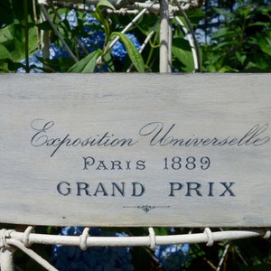 May include: A rectangular wooden sign with a distressed light gray finish featuring dark blue text that reads Exposition Universelle PARIS 1889 GRAND PRIX with a small decorative flourish centered below the final line of text.
