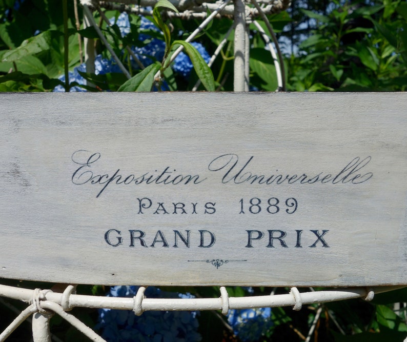 May include: A rectangular wooden sign with a distressed light gray finish featuring dark blue text that reads Exposition Universelle PARIS 1889 GRAND PRIX with a small decorative flourish centered below the final line of text.