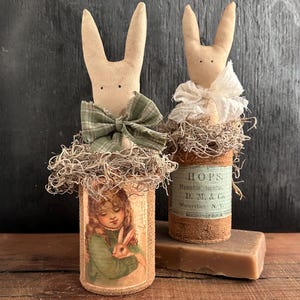 May include: Two handmade bunny figurines with fabric bodies and wire ears. The bunnies are dressed in vintage fabric and are sitting on top of repurposed glass bottles. One bottle has a label that reads "HOPS. Humulus Lupulus. D. M. & Co. Waterliet, N.Y."