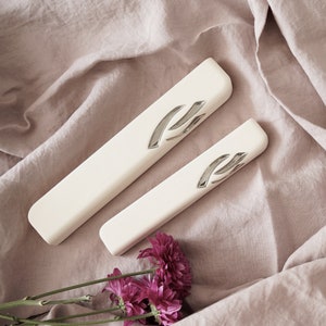 Puede incluir: Dos estuches de mezuzá blanquecinos con un diseño plateado, uno más grande que el otro, descansan sobre una tela malva. Unas ramitas de flores moradas están en la esquina inferior izquierda. Los estuches tienen forma rectangular con bordes redondeados.