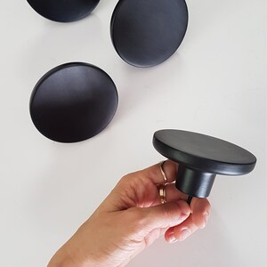 May include: Four black round wall hooks, three are flat and one is a knob with a screw. The knob is being held in a hand.