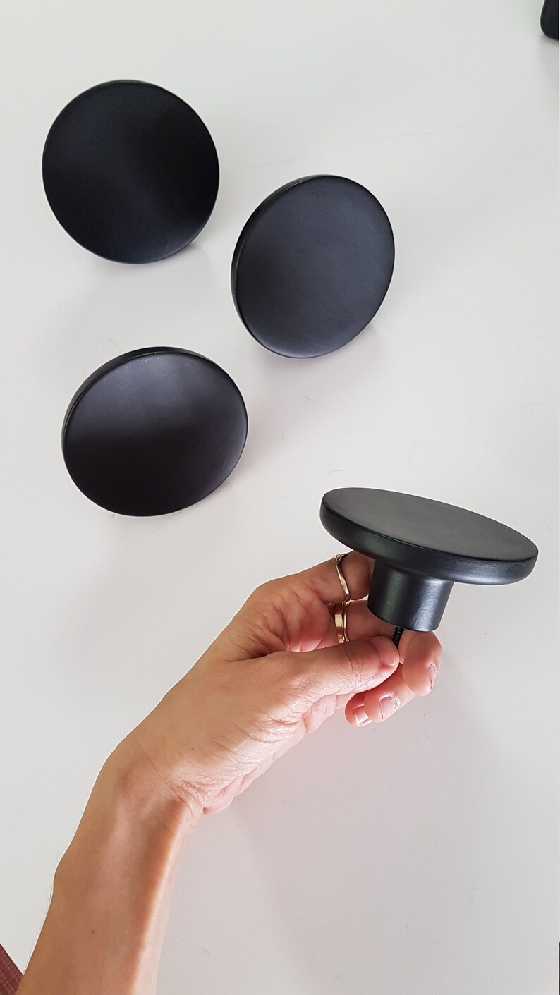 May include: Four black round wall hooks, three are flat and one is a knob with a screw. The knob is being held in a hand.