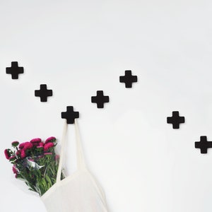 May include: A white wall with six black plus sign shaped hooks. A white tote bag is hanging from one of the hooks. A bouquet of pink flowers is in the bag.
