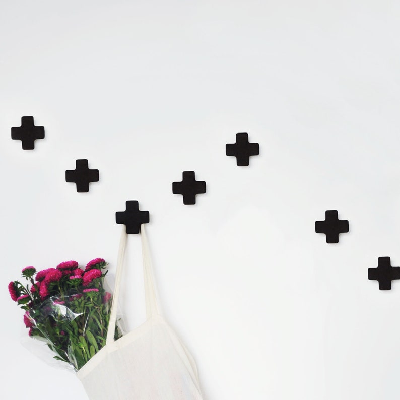 May include: A white wall with six black plus sign shaped hooks. A white tote bag is hanging from one of the hooks. A bouquet of pink flowers is in the bag.