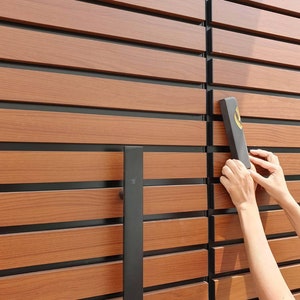 May include: A close-up of a wooden door with black horizontal slats. A black door handle is on the left side of the door. A person's hand is holding a black rectangular sign with gold lettering.