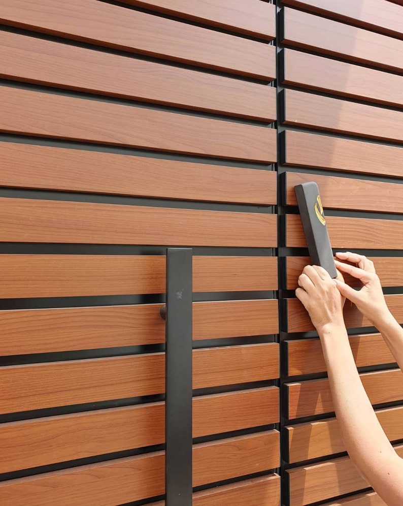 May include: A close-up of a wooden door with black horizontal slats. A black door handle is on the left side of the door. A person's hand is holding a black rectangular sign with gold lettering.