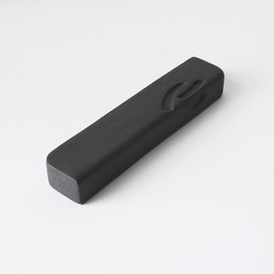Puede incluir: Un objeto rectangular negro con bordes redondeados, posiblemente una mezuzá. El objeto tiene una superficie texturizada y una letra o símbolo estilizado tallado en un extremo. El fondo es blanco liso.