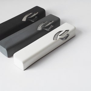 May include: Three rectangular mezuzah cases in black, gray, and white. Each case has a silver Hebrew letter Shin on the front.