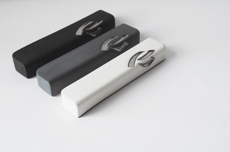 May include: Three rectangular mezuzah cases in black, gray, and white. Each case has a silver Hebrew letter Shin on the front.