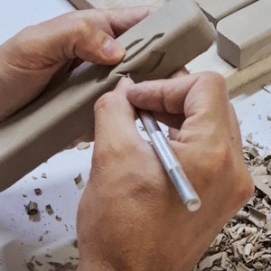 May include: A person's hands are using a small tool to carve a design into a rectangular piece of clay. The clay is light brown and the tool is silver. There are small pieces of clay on the surface around the hands.