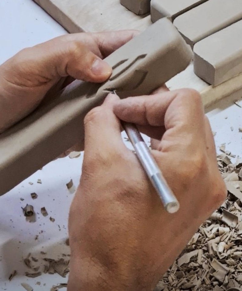 May include: A person's hands are using a small tool to carve a design into a rectangular piece of clay. The clay is light brown and the tool is silver. There are small pieces of clay on the surface around the hands.