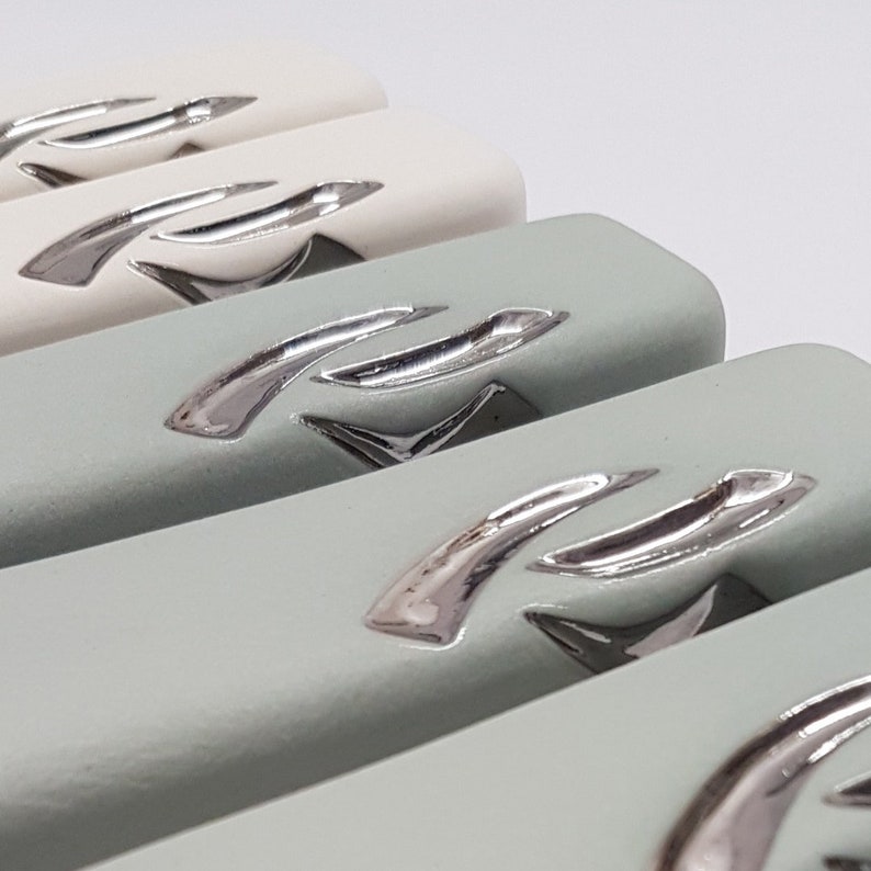 May include: Close-up of four rectangular ceramic drawer pulls with a silver metallic finish. The pulls have a curved design with a raised, stylized letter 'R' on each.