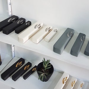 May include: A shelf display of various mezuzahs in different colors and sizes. The mezuzahs are made of wood and have a silver or gold Hebrew letter Shin on the front. The mezuzahs are arranged in rows on a white shelf.