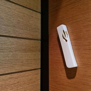 May include: A white mezuzah with a gold Hebrew letter Shin on a wood panel door.