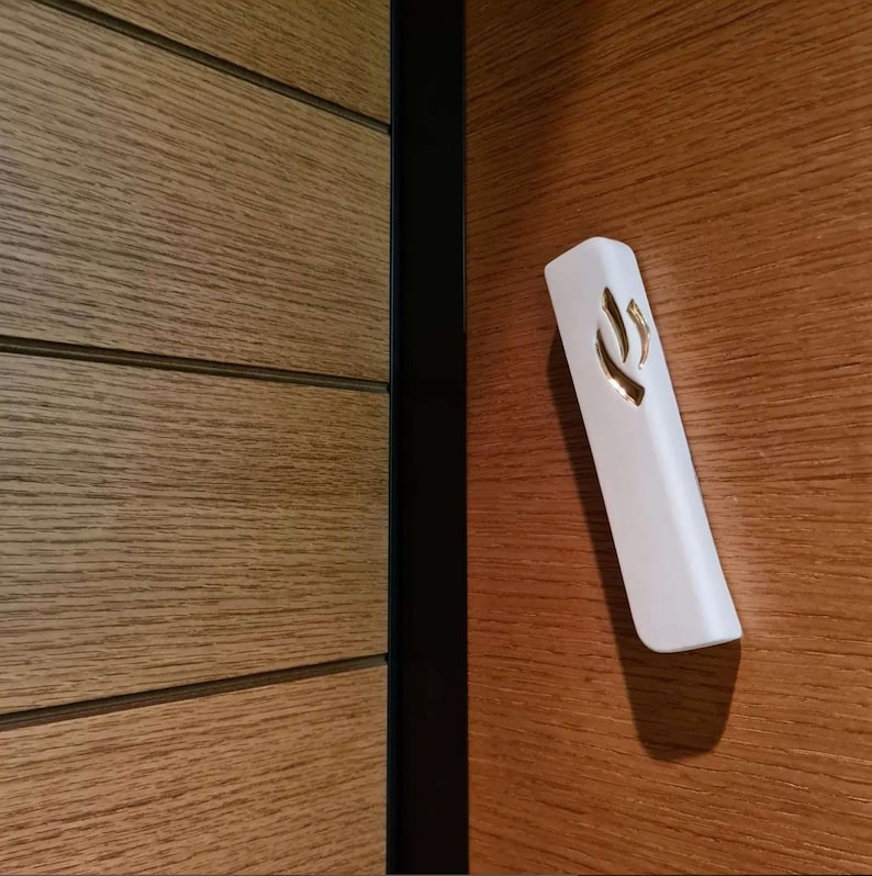 May include: A white mezuzah with a gold Hebrew letter Shin on a wood panel door.