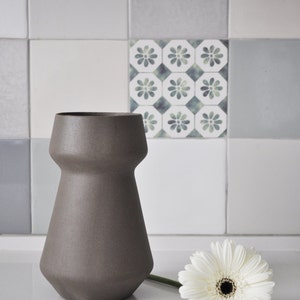 May include: A gray ceramic vase with a unique shape, paired with a white gerbera daisy. The vase has a wide base and a narrower neck. The background features a tiled wall with various shades of gray and white.