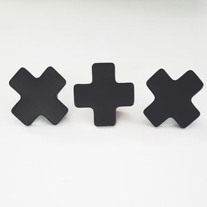 May include: Three black metal wall hooks in the shape of a plus sign and a cross. The hooks are mounted on a white wall.
