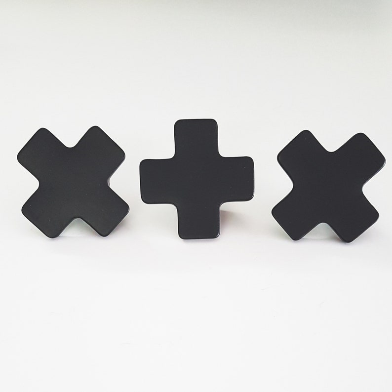 May include: Three black metal wall hooks in the shape of a plus sign and a cross. The hooks are mounted on a white wall.
