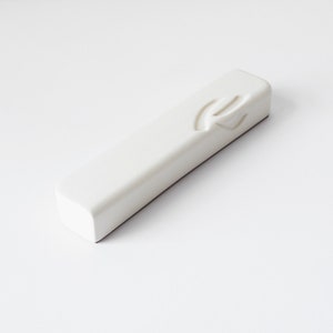 Puede incluir: Una caja de mezuzá rectangular blanca con una letra hebrea Shin tallada en la parte delantera.
