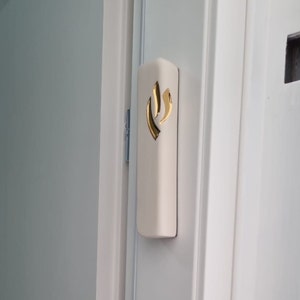 Op de afbeelding: Een witte mezuzah met een gouden vlamontwerp. De mezuzah is bevestigd aan een wit deurkozijn.