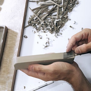 May include: A person's hand is using a tool to trim a piece of clay. The clay is light brown and has a rough texture. There are small pieces of clay on the white surface.