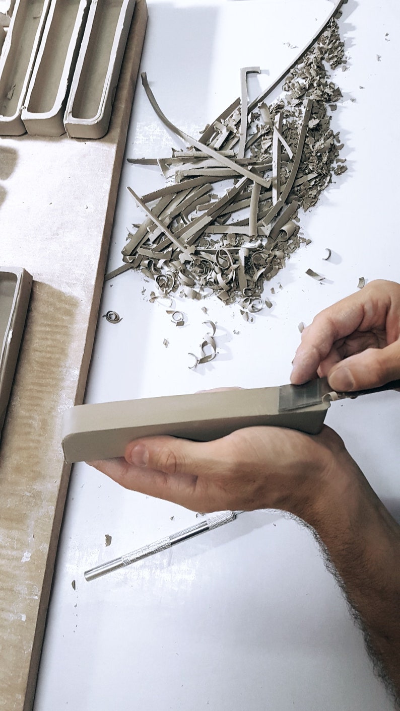 May include: A person's hand is using a tool to trim a piece of clay. The clay is light brown and has a rough texture. There are small pieces of clay on the white surface.