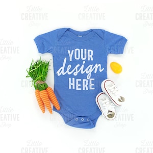 May include: A blue baby onesie with the text "YOUR design HERE" in white. The onesie is laid flat on a white background with a bunch of carrots, a yellow toy, and a pair of white baby shoes.