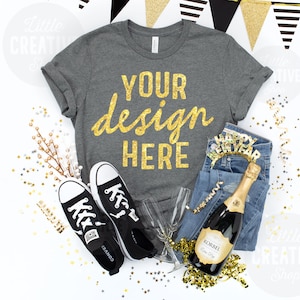 May include: A flat lay featuring a gray t-shirt with gold text reading "YOUR design HERE", black Converse sneakers, blue jeans, champagne, and a "Happy New Year" headband. Gold confetti and decorative banners complete the festive scene.
