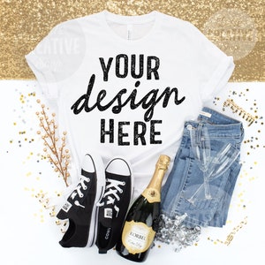 May include: A white t-shirt with the text "YOUR design HERE" is paired with blue jeans, black sneakers, a champagne bottle, and champagne flutes. The items are arranged on a white surface with gold glitter and star-shaped confetti.