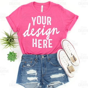 May include: A vibrant pink t-shirt with the text "YOUR design HERE" in white script is styled with denim shorts and white trainers. A small succulent and a green plant are placed near the shirt. The t-shirt is rolled up at the sleeves.