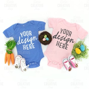 May include: Two baby onesies, one blue and one pink, with the text "Your design here" on each. The onesies are surrounded by Easter-themed decorations, including a nest with eggs, carrots, and green grass.