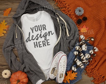 May include: A white t-shirt with the text "YOUR design HERE" on it, laying on a gray hoodie, blue jeans, and a brown blanket. There are also fall-themed decorations like pumpkins, leaves, and cotton bolls.