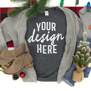 以下が含まれることがあります： 白い文字で「YOUR design HERE」と書かれたグレーのTシャツ。Tシャツは、グレーのカーディガン、茶色のブーツ、青いジーンズ、小さな松の木、赤と黒のチェック柄の毛布と一緒に白い表面に置かれています。