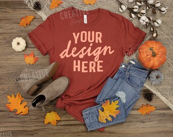May include: A rust-colored t-shirt with the text "YOUR design HERE" in a light peach color. The t-shirt is laid out on a wood surface with fall leaves, a pumpkin, and a pair of gray ankle boots.