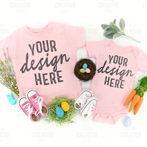 May include: Two pink t-shirts with the text "Your design here" on them. The shirts are laid out on a white background with Easter decorations, including a nest with eggs, carrots, and flowers.
