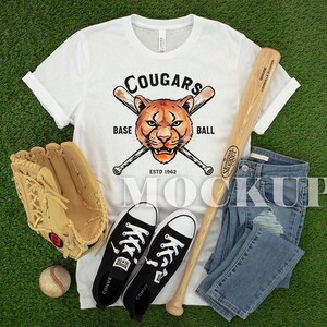 May include: White t-shirt with a cougar head and baseball bats graphic, baseball bat, baseball glove, baseball, black Converse trainers, and blue jeans on a green surface. The shirt says "Cougars Base Ball Estd 1962".