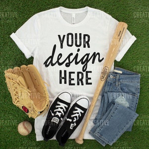 May include: A white t-shirt with the text "YOUR design HERE" in black, alongside a baseball bat, glove, ball, blue jeans, and black Converse trainers. The items are arranged on a green surface.