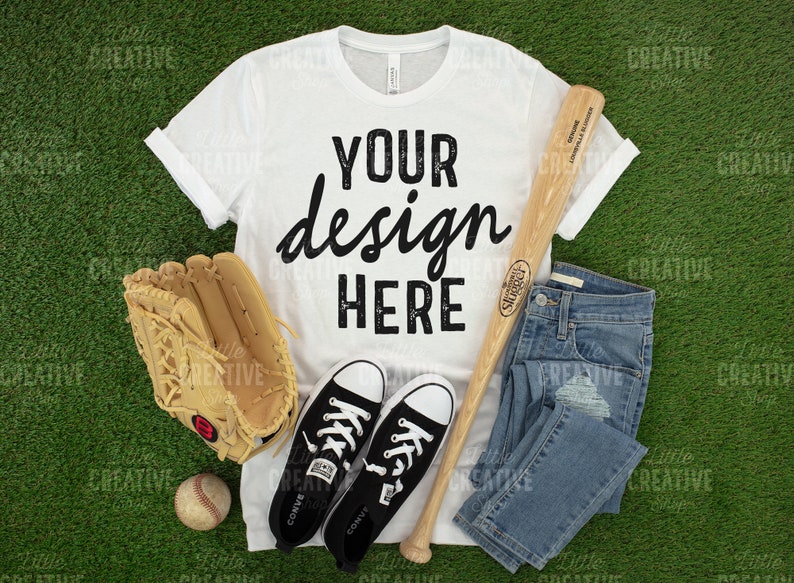 May include: A white t-shirt with the text "YOUR design HERE" in black, alongside a baseball bat, glove, ball, blue jeans, and black Converse trainers. The items are arranged on a green surface.