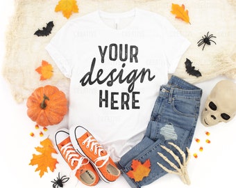 May include: White t-shirt with the text "YOUR design HERE" in black. The shirt is lying flat on a white surface with a pair of orange trainers, a pumpkin, autumn leaves, a skeleton hand, a skull, and a pair of blue jeans.