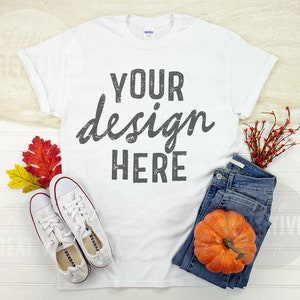 May include: White t-shirt with the text "YOUR design HERE" in a distressed font. The shirt is laid flat on a white wooden surface with a pair of white sneakers, a pair of blue jeans, a small pumpkin, and fall foliage.