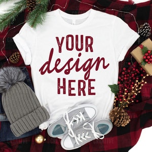 May include: White t-shirt with the text "YOUR design HERE" in maroon lettering on a red and black plaid blanket with a gray beanie, pine cones, and red berries.